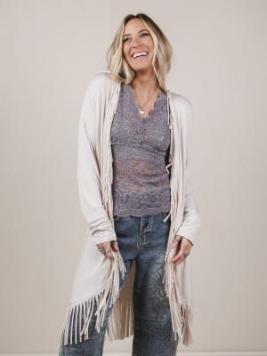 Woman wearing a long beige cardigan with fringe, purple top, and jeans against a plain background