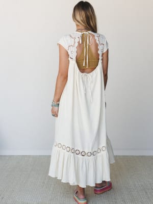 Shot from behind, a cream maxi dress with an open back and delicate eyelet detailing is styled with a mustard crochet bralette peeking through, layered boho jewelry, and rainbow platform slides — effortlessly free-spirited and ready for warm weather.