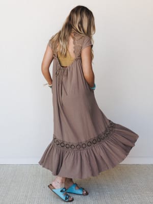 Shot from the back, a flowy mocha maxi dress fans out mid-twirl, showing off its open lace back, crochet hem band, and ruffled skirt — styled with a mustard bralette peeking through and turquoise sandals for that effortlessly layered, lived-in boho f