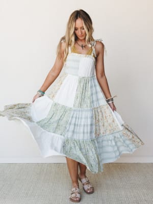 A breezy patchwork midi dress in ivory, sage, and soft floral panels swirls mid-movement, styled with slide sandals and stacked bracelets against a clean studio wall — the kind of dress you wear when you want to feel like you're already somewhere bea