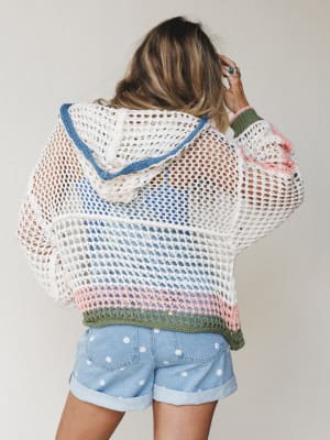 Shot from the back, she's wearing an open-weave crochet hoodie in ivory with soft pastel stripe details — blue, blush pink, and olive — layered over polka dot denim shorts, hand tucked into her hair like she's mid-thought on a breezy afternoon.