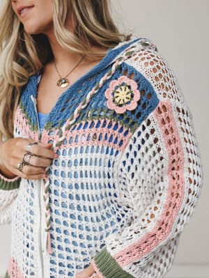 A close-up of a multicolor crochet zip-up jacket layered over a blue tank, with a granny square chest panel, 3D floral appliqué, and colorblock striping — the kind of piece that makes a Tuesday feel like a road trip.