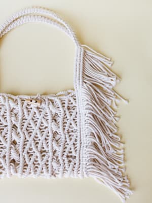 A close-up of a macrame tote bag in soft ivory, shot flat against a warm cream background — the knotted texture and fringe trim do all the talking.