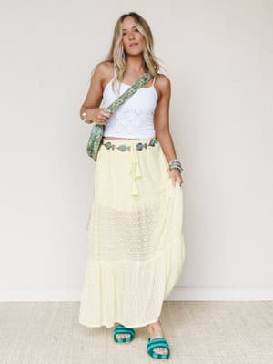 A sun-warmed outfit built around a pale yellow maxi skirt and white eyelet crop tank, styled with a beaded fish chain belt, teal crossbody bag, and turquoise slide sandals — easy, summery, and just a little playful.