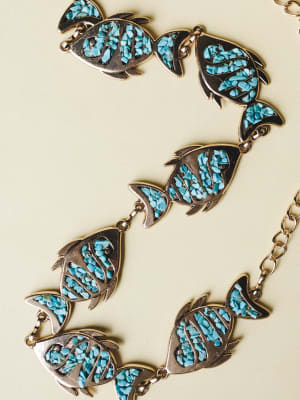 A vintage-inspired chain belt laid flat, featuring linked fish-shaped medallions filled with crushed turquoise stone set in gold-toned metal — earthy, a little wild, the kind of piece that makes an outfit feel like a story.