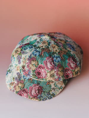 A tapestry-style newsboy cap in a richly colored floral print, shot against a warm dusty rose backdrop — the kind of hat that makes a plain outfit feel like a whole personality.