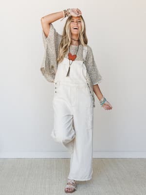 A laughing model in wide-leg ivory overalls layered under an open-knit oversized cardigan, styled with bold boho jewelry — the kind of outfit that looks like it was thrown on and somehow landed perfectly.