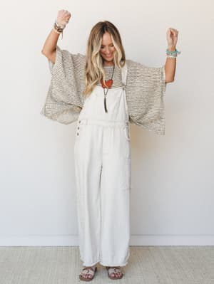 A smiling model with arms raised joyfully wears wide-leg ivory overalls layered under a flowy open-weave kimono top, styled with stacked bracelets, a long pendant necklace, and sandals — the whole look has that effortless, happy-to-be-here energy.