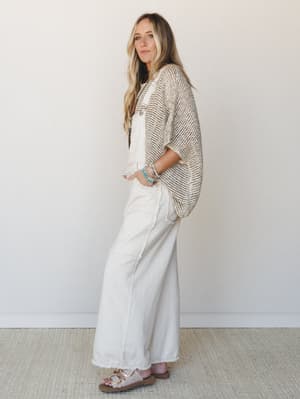 A relaxed full-body shot of a model in cream wide-leg overalls layered under an open-knit sweater top, styled with stacked turquoise bracelets and birkenstock-style sandals — the kind of effortless outfit that somehow looks like it took no effort at