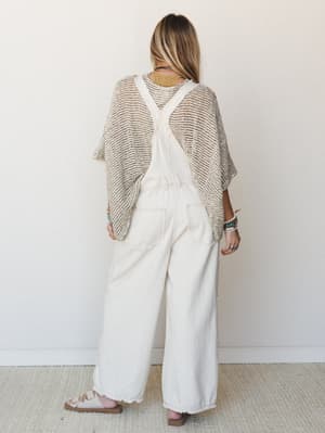 Shot from the back, a model layers an open-knit poncho over wide-leg ivory overalls, styled with stacked turquoise bracelets and strappy sandals — the kind of effortless, collected outfit that looks like it came together naturally.