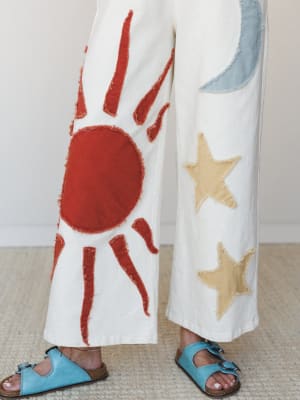 A close-up of wide-leg ivory pants covered in raw-edge fabric appliqué — a bold sun on one leg, crescent moon and stars on the other — paired with turquoise footbed sandals on a woven rug.