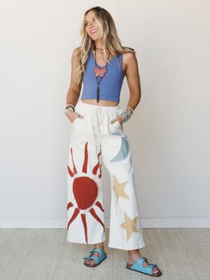 A smiling model pairs statement ivory wide-leg pants — patched with a bold red sun, gold stars, and a blue moon — with a periwinkle ribbed crop tank and turquoise sandals, giving the whole look a free-spirited, celestial-art-project energy.