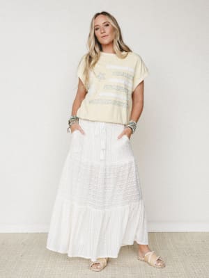 A breezy, sun-washed look — cream tiered maxi skirt with a textured stripe detail paired with a butter yellow graphic tee, stacked turquoise bracelets, and slide sandals, shot clean against a soft white studio wall.