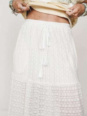 A close-up detail shot of a cream lace tiered maxi skirt with a drawstring tassel waist, styled with a soft yellow cropped top and stacked turquoise bracelets — effortlessly soft and boho all the way through.