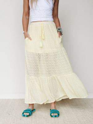 A breezy butter-yellow tiered maxi skirt with a sheer lace panel and tassel drawstring waist, styled with a white eyelet crop top and teal crochet sandals — the kind of outfit that looks like it was assembled effortlessly on a warm morning.