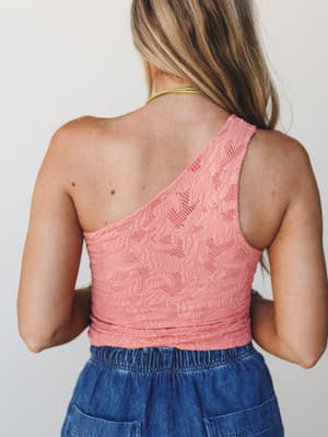 Shot from the back, a model in a coral-pink one-shoulder lace brami top paired with a denim skirt — the open-back detail and textured floral lace do all the talking.