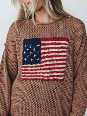 An open-knit caramel sweater with a bold American flag graphic knitted into the chest — the kind of piece that reads patriotic without trying too hard, lived-in and relaxed in all the right ways.