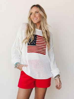 A smiling blonde model wearing an oversized open-knit white sweater with an American flag graphic on the chest, paired with bright red shorts and stacked beaded bracelets — casual, sun-soaked, and ready for a backyard cookout.