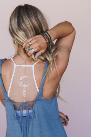 A woman with long blonde hair in a blue sleeveless top reveals the Crescent Moon Tattoo Bralette - White and a temporary crescent moon floral tattoo on her upper back, accessorized with rings and bracelets as she touches her hair.