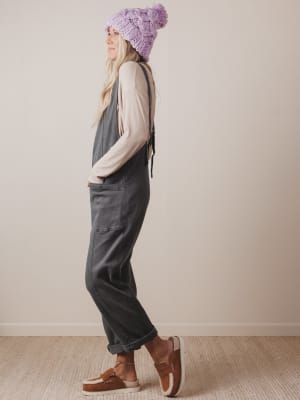 Person wearing a purple knit hat, gray overalls, and brown shoes against a plain background