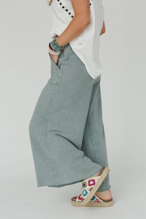 A person stands sideways wearing the Relaxing Robin Wide Leg Pant in Faded Teal, a white short-sleeve top, colorful beaded bracelets, and espadrille sandals with a multicolored crochet pattern.