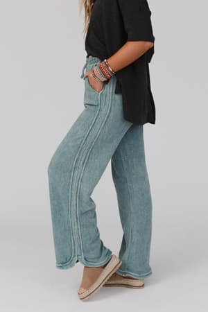 A person stands sideways in the So Comfy Wide Leg Pant Full Length - Teal, paired with a loose black top, beige platform sandals, and colorful beaded bracelets. The background is plain and light.