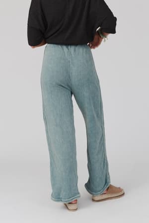A person faces away, wearing So Comfy Wide Leg Pant Full Length - Teal with a drawstring waist, a black rolled-sleeve top, and beige sandals against a plain light background.