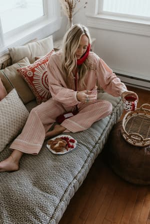 A soft morning moment — model sits cross-legged on a boho daybed in a matching pink gingham pajama set, holding a mug and picking at a plate of raspberries, the whole scene radiating slow-morning, stay-in-your-pajamas energy.
