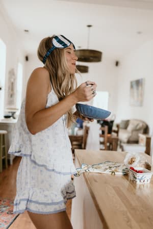 A relaxed morning moment — model stands at a kitchen counter in a soft blue floral PJ short set, sleep mask pushed up on her head, eating from a bowl like she has absolutely nowhere to be.