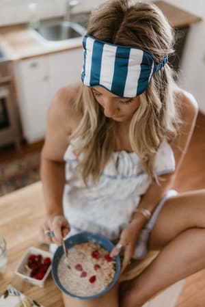 A blonde model sits cross-legged on a kitchen counter eating a bowl of oatmeal with berries, wearing a blue and white striped sleep mask pushed up on her forehead and a floral sleeveless top — the whole vibe is slow morning, no apologies.