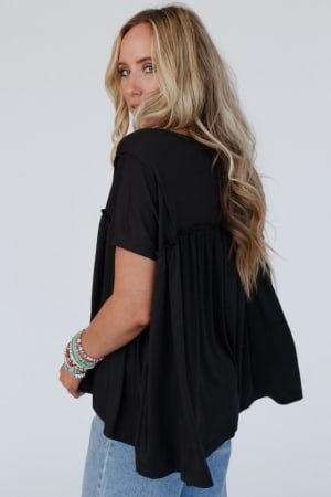 A woman with long, wavy blonde hair wears the Sunday Morning Babydoll Top - Black with light blue jeans and colorful beaded bracelets. She looks over her shoulder against a plain white background.