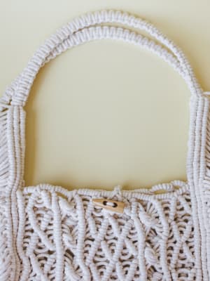 A close-up of a cream macramé tote bag with intricate knotted texture and a natural wood toggle button closure — the kind of bag that looks handmade in the best possible way.