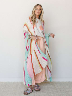 A bright, playful layered look built around a floor-length open-knit mesh duster in a multicolor stripe pattern, worn over a seashell graphic tee and a soft peach maxi skirt — the whole outfit has that easy, colorful energy of a beach town afternoon.