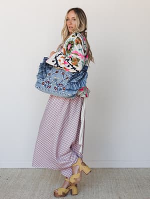 A model glances back over her shoulder in a richly layered boho outfit — bold floral embroidered top, soft mauve geometric midi skirt, and a ruffle-trimmed embroidered denim tote, finished with studded mustard platform sandals against a clean studio