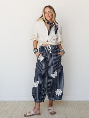 A relaxed full-body model shot featuring navy striped balloon pants with crochet butterfly and floral appliqués, styled with a knotted cream button-down, navy bandana necktie, stacked bracelets, and white sandals — easy, expressive, and just the righ