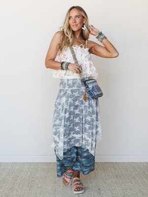 A layered boho look full of texture and personality — a floral strapless ruffle top paired over a lace overlay midi skirt with camo wide-leg pants peeking beneath, styled with stacked bracelets, a beaded crossbody bag, and platform sandals against a