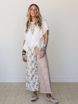 A white lace poncho top paired with mixed-print wide-leg pants — one leg floral, one leg leopard — styled with stacked beaded bracelets and woven flats for a free-spirited, effortlessly layered look.