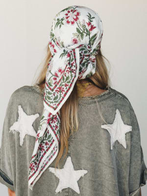 Shot from behind, a model wears a floral block-print bandana tied over her head, trailing down her back alongside loose blonde hair, paired with an oversized washed grey tee featuring distressed white star appliqués — the kind of outfit that looks ef