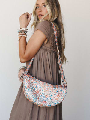 A sun-kissed model glances back over her shoulder wearing a flowy taupe maxi dress with lace back detail, styled with a crescent-shaped floral quilted crossbody bag and layers of mixed beaded bracelets — the whole look feels like a good Saturday with