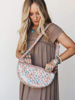 A model in a flowy taupe maxi dress looks down as she opens a crescent-shaped quilted crossbody bag in a coral, blue, and cream floral print — stacked rings and turquoise jewelry completing the relaxed boho moment.