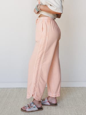 A side-profile shot of wide-leg peach pants with contrast stitching, styled with a cropped white tee, beaded bracelets, and embellished sandals — relaxed and ready for wherever the day goes.