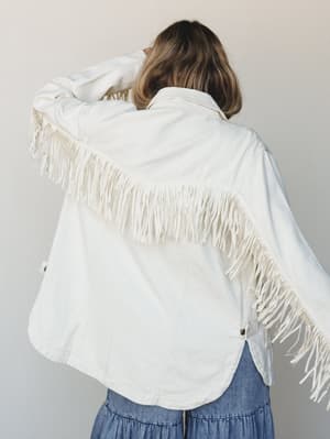 Shot from behind, a model shows off the dramatic fringe detail across the back yoke of a cream denim jacket, paired with a tiered chambray midi skirt — the kind of outfit that looks just as good at a concert as it does on a Sunday.