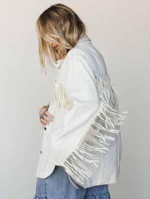A side profile shot of a model in an oversized white denim jacket with dramatic fringe trim down the sleeves and back, styled with a blue denim skirt and turquoise ring — effortlessly boho with a Western edge.