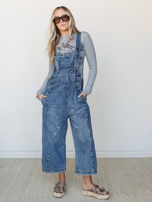 Wide-leg denim overalls with a lived-in wash and distressed detailing, styled over a long-sleeve graphic tee with layered gold necklaces and leopard print platform sandals — the kind of outfit that looks effortless but totally isn't.