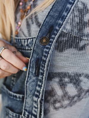 A close-up detail shot highlighting the branded brass snap button on acid wash denim overalls worn over a printed knit top, with stacked beaded necklaces and painted nails peeking into frame — the kind of layered, lived-in outfit that looks effortles