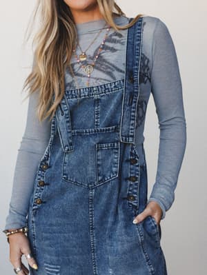 A denim overall dress layered over a sheer gray long-sleeve printed top, styled with layered boho necklaces and stacked jewelry — the kind of outfit that looks effortless but clearly had a vision.
