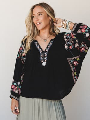 A relaxed black dolman-sleeve top covered in vibrant floral embroidery styled with a sage pleated skirt and layered jewelry — the kind of piece that does all the talking so you don't have to.