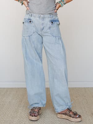 Light wash wide-leg denim pants with cargo-style flap pockets and drawstring ankles, styled casually with a grey tee, stacked turquoise bracelets, and leopard print platform sandals — relaxed and a little lived-in, in the best way.