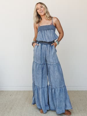 A wide-leg chambray jumpsuit with tiered, flowy legs and smocked bodice and waist styling — worn with stacked turquoise bracelets and sandals, it's the kind of outfit that looks effortless but clearly wasn't an accident.