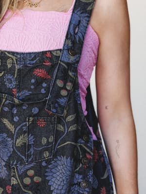 A close-up of floral-printed black denim overalls layered over a pink textured tank, with brass snap hardware and a peek of a gold 'MAMA' necklace — the kind of outfit that's doing a lot quietly.
